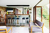 Gepolsterte Sitzbank im 50er Jahre Stil in Loungebereich, im Hintergrund Retro Barhocker vor Theke in offenem Wohnraum, seitlich Glasfalttür zum Garten