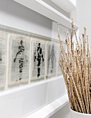 Getrocknete Stengel in Vase vor Wand mit Zeichnungen auf bedruckten Buchseiten in weissen Bilderrahmen