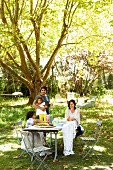 Familie in sonnenbeschienenem Garten um gedecktem Tisch unter schattigem Baum in sommerlicher Stimmung