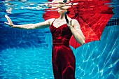 A woman under water in a swimming pool wearing a red dress, holding a red umbrella