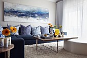 Gemälde mit Meermotiv über Sofa mit blauen Kissen, davor schlichte Holztische