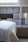 Schlafzimmer in eleganten Grautönen, auf Bett glänzende Tagesdecke vor gepolsterter Kassettenverkleidung an Wand, seitlich Nachttisch mit Tischleuchte