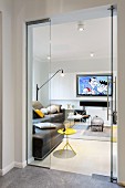 View through open glass doors of yellow side table, brown leather sofa and flatscreen TV on wall