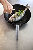 A fish larded with herbs and lemons being drizzled with oil in a pan