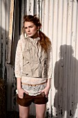 A young woman wearing leather shorts, a lace blouse and a leather gillet standing in front of a corrugated iron wall