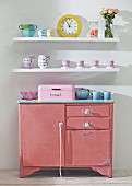 Battered pink kitchen cabinet below crockery on white floating shelves