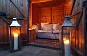 Large, lit candles in floor-standing candle lanterns outside wooden cabin with sofa in illuminated interior
