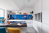 A breakfast bar in a white designer kitchen with a blue glass panel on the wall with a built-in touch screen