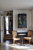 Two antique chairs around bistro table and small display cabinet above radiator next to open door with view into hallway