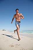 Junger Mann mit freiem Oberkörper und Badeshorts joggt am Strand