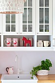Mugs with reindeer motif below glass-fronted wall units above white sink and potted herbs