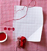 Weißes Blatt Papier auf rot-weiß kariertem Stoff mit verschiedenen Garnrollen und roter Farbe