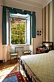 Ausschnitt eines Schlafzimmers, Blick über Bett auf offenes Fenster mit lässig drapiertem Vorhang über Vorhangstange