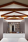 Renoviertes Schlafzimmer mit sichtbarer rustikaler Tragwerkskonstruktion, Blick über Bettkopfteil auf hellgraue Schränke und Bücherwand