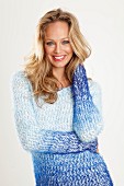 A blonde woman wearing a blue-toned, coarse-knit jumper with her hand on her hair