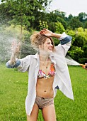 A woman wearing colourful underwear and an open man's shirt being sprayed with a hose