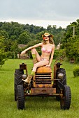 A woman wearing underwear and a straw hat sitting on an old tractor with a flower in her mouth