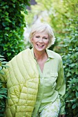Ältere Frau mit hellgrüner Bluse, grün-weisser Hose und Steppjacke im Garten