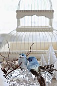 Two bird ornaments on crystal bowl in front of vintage bird cage