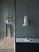 Table lamp on bedside cabinet against exposed concrete wall and vase of exotic flowers on small metal table in background