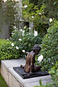 Bronzeskulptur auf niedriger Mauer im Garten; weiße Rosen im Hintergrund