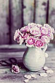Ein Strauß rosa Rosen in Keramikkrug, daneben einzelne Blüte und Blütenblätter