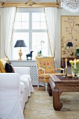 White sofa, antique solid-wood coffee table and armchair below window with white, draped curtains in living room