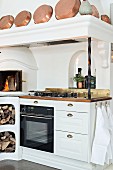 weiße Küchenzeile im Landhausstil mit Backofen, Kamin, Holzlager und Kupferpfannen auf Sims der Abzugshaube