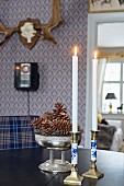 Candlesticks and antique silver bowl of pine cones on table in front of hunting trophy on blue wallpaper