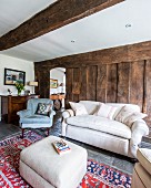 Rustikales Wohnzimmer mit hellem Polsterhocker vor Couch und Sessel, im Hintergrund Holzwand aus Bohlen