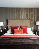Scatter cushions arranged on double bed with tall headboard against wallpaper with pattern of birch trunks
