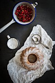 A Bundt cake dusted with icing sugar on a piece of white paper with redcurrants in a vintage sieve on a grey slate surface