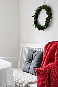 Grey felt cushion and red and white patterned blanket on white-painted bench below Christmas wreath on wall