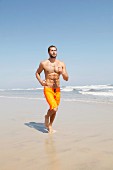 A muscular young man wearing yellow Bermuda shorts jogging along a beach