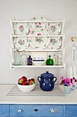 Detail of fruit bowl and blue ceramic pot on kitchen counter below decorative wall plates in plate rack mounted on wallpapered wall