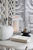 Halloween arrangement of white-painted pumpkin, skull ornament under glass cover and romantic candle lantern on table