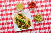 Vegan tacos with kale, guacamole, salsa, and salsa verde on a red-and-white checked tablecloth