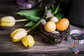 Osternest mit Eiern, buntem Geschenkband und gelben Tulpen auf rustikaler Holzunterlage