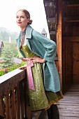 A woman wearing a green dirndl and a short coat standing on the balcony of a farm house
