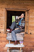 A woman wearing a patterned woollen jacket and checked trouser sitting in an open window of a wooden house
