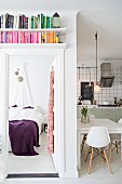 Bücherregal über offener Schlafzimmertür und Blick auf Bett mit violetter Tagesdecke, seitlich offene Küche mit Essplatz