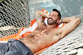 A laughing, topless man wearing shorts lying in a hammock