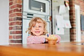 A boy standing at a kitchen table with a muffin in front of him