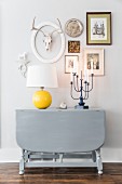 Table lamp with yellow base on grey-painted, drop-leaf console table below pictures and ornaments on wall
