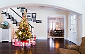 Red and white wrapped gifts under Christmas tree decorated with fairy lights in spacious hallway of American country house