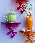 Brightly coloured wall brackets holding orange vase, orchid sprig and cactus in vintage teacup