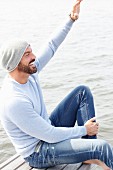 A man wearing a grey knitted hat, a light-blue jumper and jeans by the sea