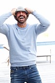 Mann mit Vollbart in hellblauem Pullover und Jeans macht Spaß mit Strickmütze