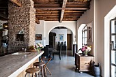 View past kitchen counter into open-plan living area with arched open doorway in renovated country house with rustic wood-beamed ceiling