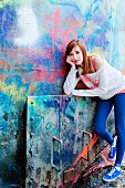 A young woman standing against a colourfully sprayed wall looking bored